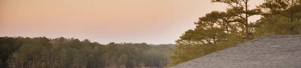 A picturesque scene at sunset with a gray-roofed building partially visible in the foreground. The background features dense green trees and a soft pinkish-orange sky, evoking a tranquil, rural setting.