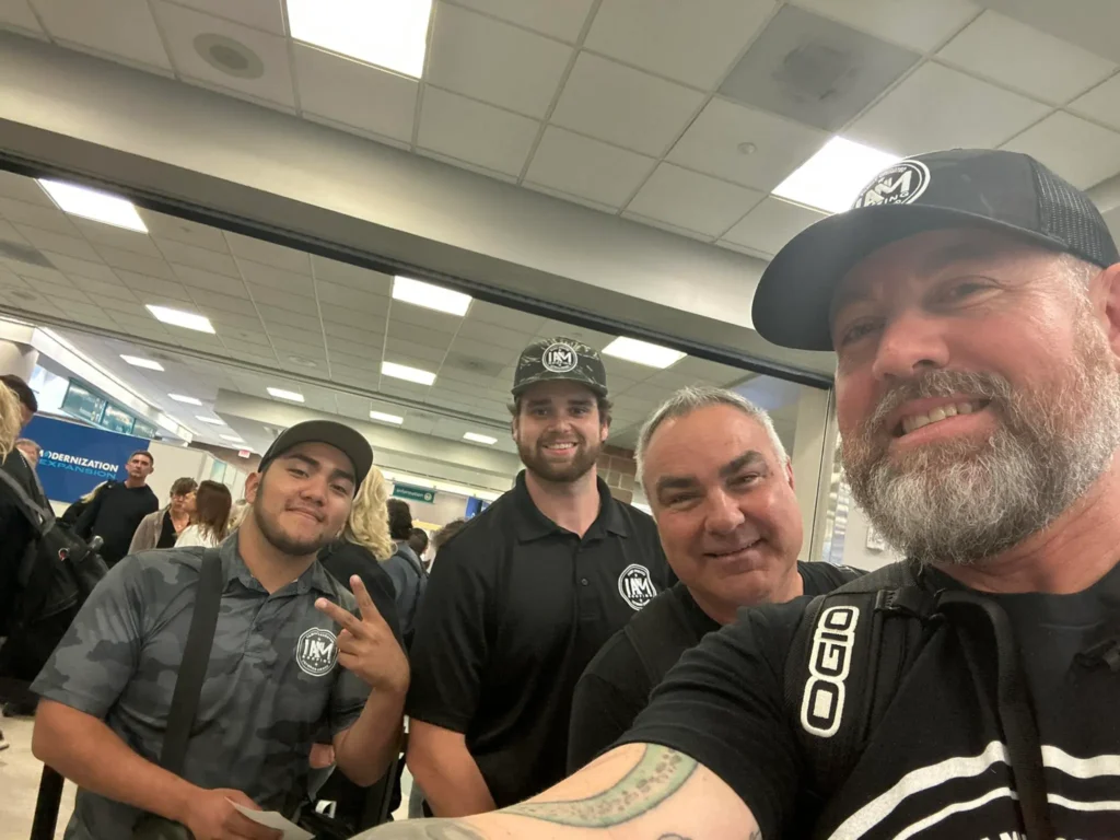 Four men in casual attire, smiling together at an airport terminal. Two of them wear black caps and shirts with a logo. One man is making a peace sign. The background shows people and a sign for Continental Express.