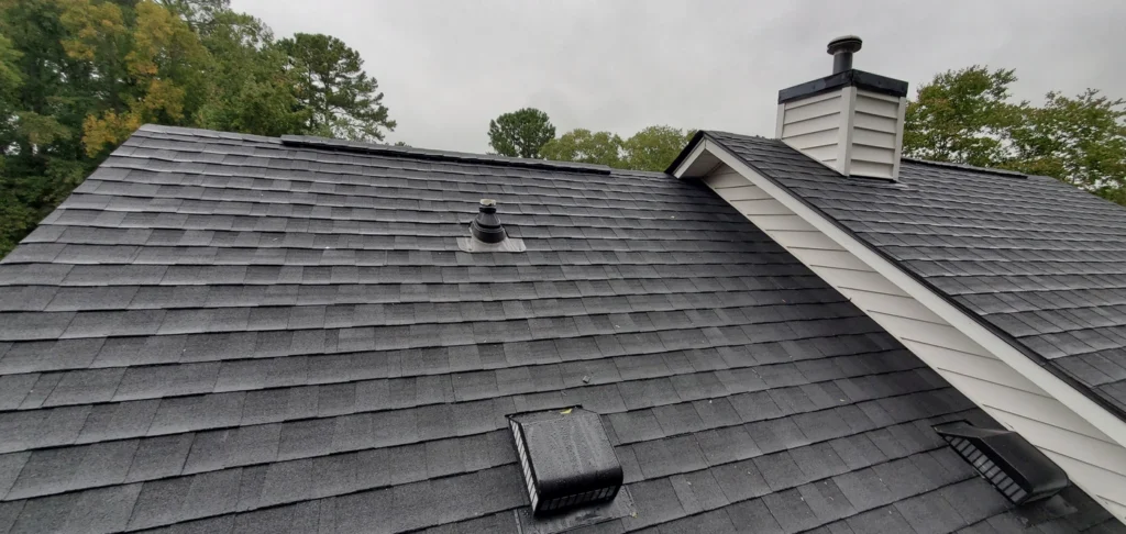 A sloped roof with gray asphalt shingles showcases a discreet roof patch, along with a chimney, vent pipes, and a roof vent. Trees stand tall in the background under a cloudy sky.