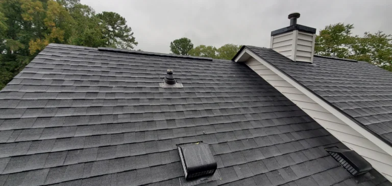 A sloped roof with gray asphalt shingles showcases a discreet roof patch, along with a chimney, vent pipes, and a roof vent. Trees stand tall in the background under a cloudy sky.