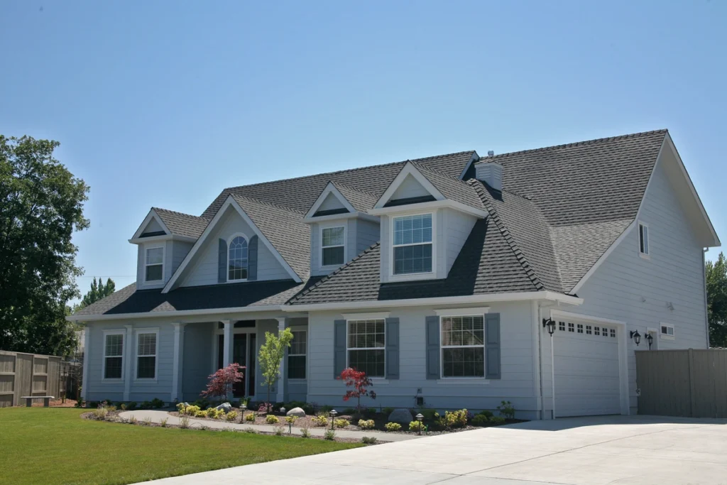 A large two-story house with a steep gable roof, featuring light gray siding and multiple windows. The metal roof, preferred over shingles for durability, complements the manicured front yard with small shrubs and trees. A wide driveway leads to a garage on the right, under a clear blue sky.